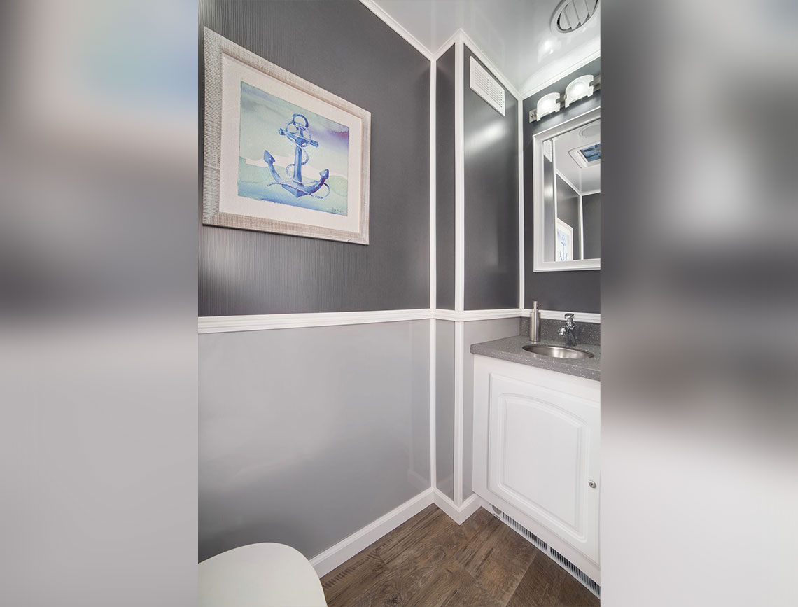 Restroom Trailer featuring a modern interior with gray walls, stylish white trim, and a framed watercolor painting of an anchor. It includes a sleek gray countertop with a sink, a mirror, and wooden flooring.