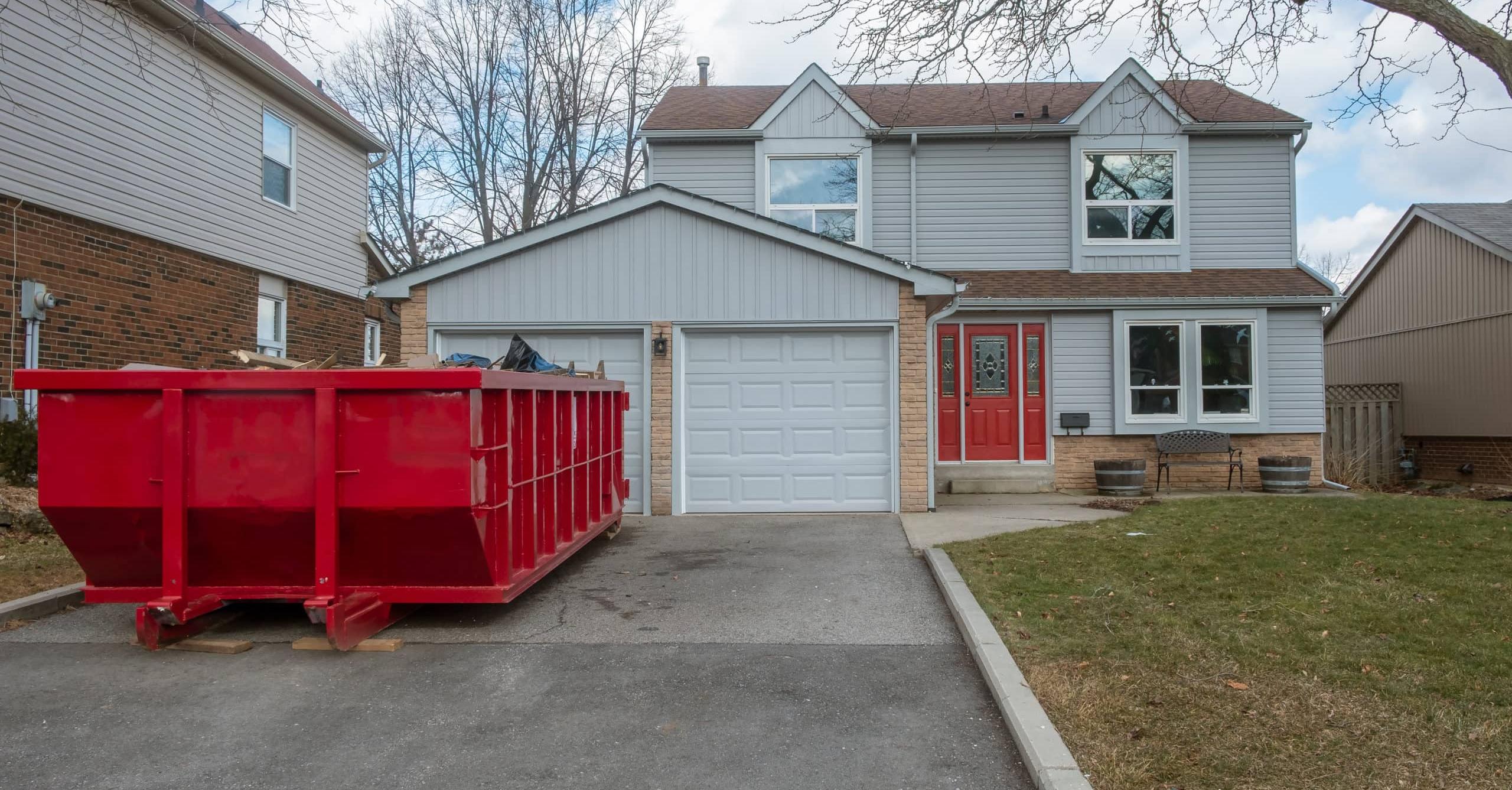 A large red roll off dumpster is positioned in front of a two-story house with gray siding and a gabled roof. The house features red doors and large windows. The driveway is paved, with some grass and brown leaves visible on the lawn. Trees are bare in the background, indicating early spring.